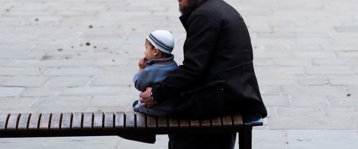 An adult and a small child in a striped hat sitting together on a slatted bench beside a paved walk