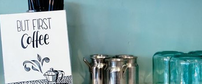 A cafe counter with steel milk jugs and stacked glassware against a painted wall, a printed card beside them reading But First Coffee
