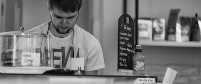 A man working behind a cafe counter beside a domed cake stand labelled Victoria Sponge, with a chalkboard menu and retail coffee bags on the shelf behind