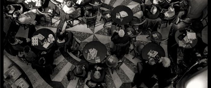 Round cafe tables photographed from above on a patterned stone floor, customers seated around them with newspapers